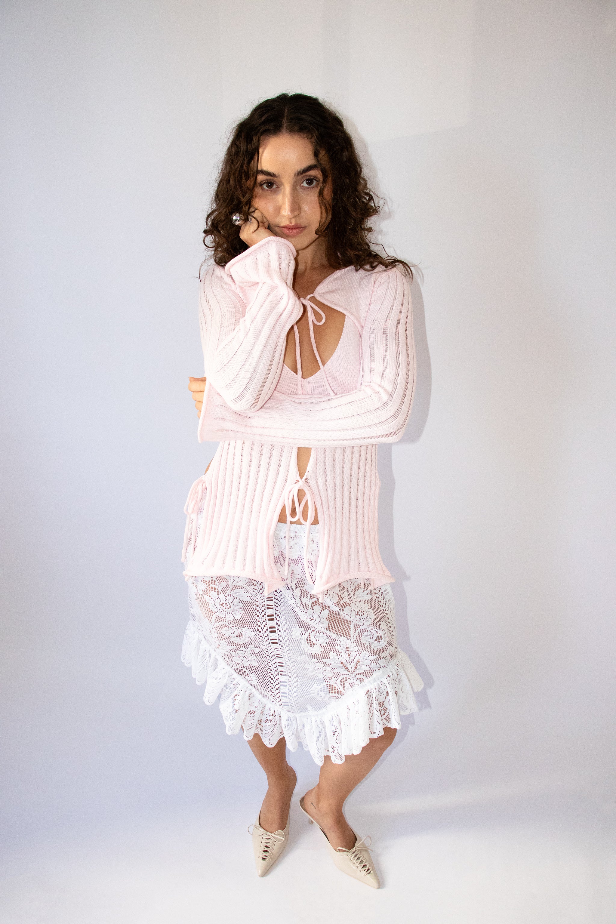 Woman wearing a light pink knit top and bolero with a white lace skirt on a white background